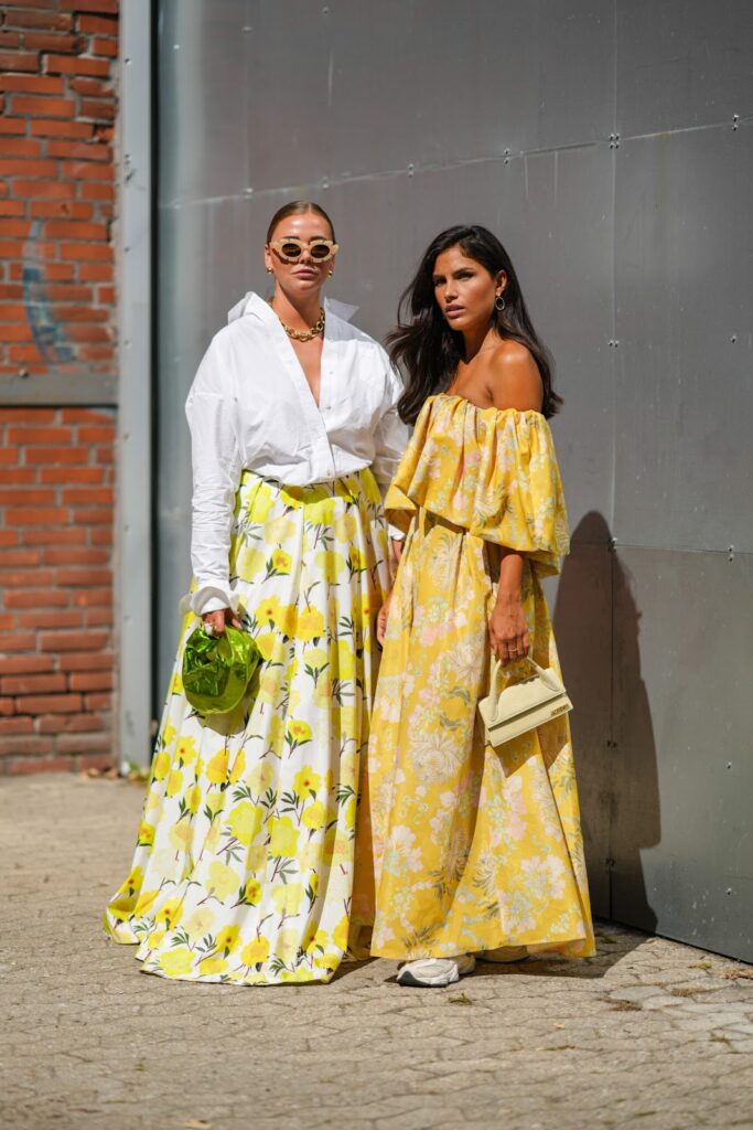 Kobiety ubrane we wiosenne stylizacje - białą oversizową koszulę wpuszczoną w kwiecistą spódnicę maxi oraz żółtą sukienkę w kwiecisty wzór i sneakersy