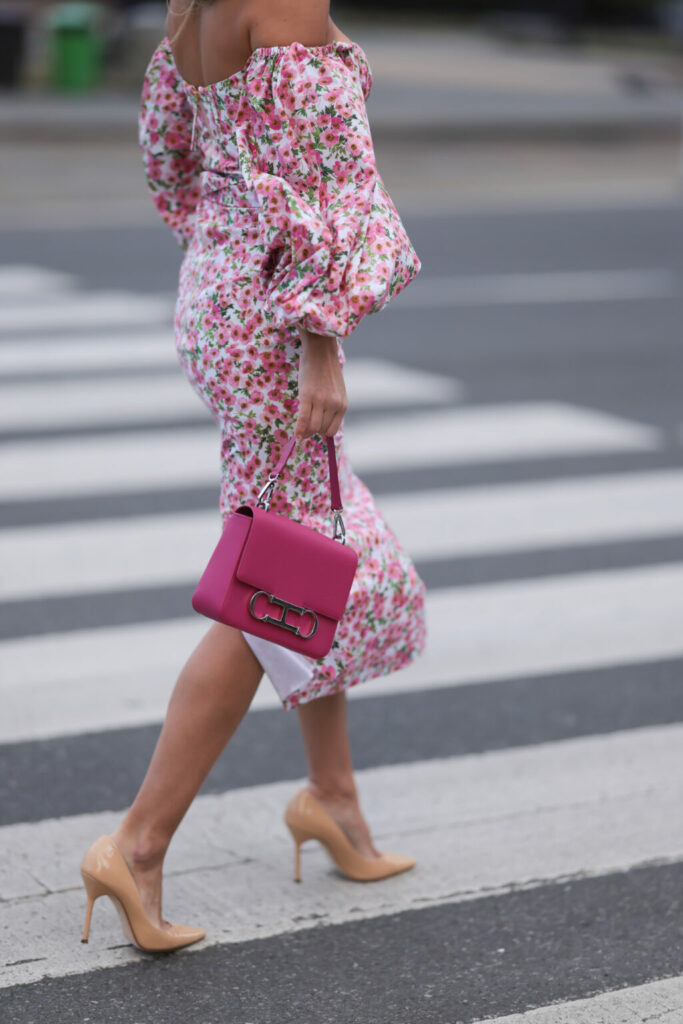 Kobieta w kwiecistej sukience z bufiastymi rękawami odsłaniającymi ramiona z torebką w intensywnym różowym kolorze i beżowych lakierowanych szpilkach